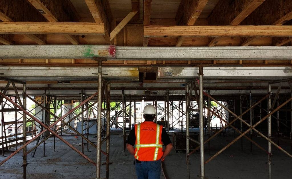 A man stands beneath a wood structure, surveying concrete forming